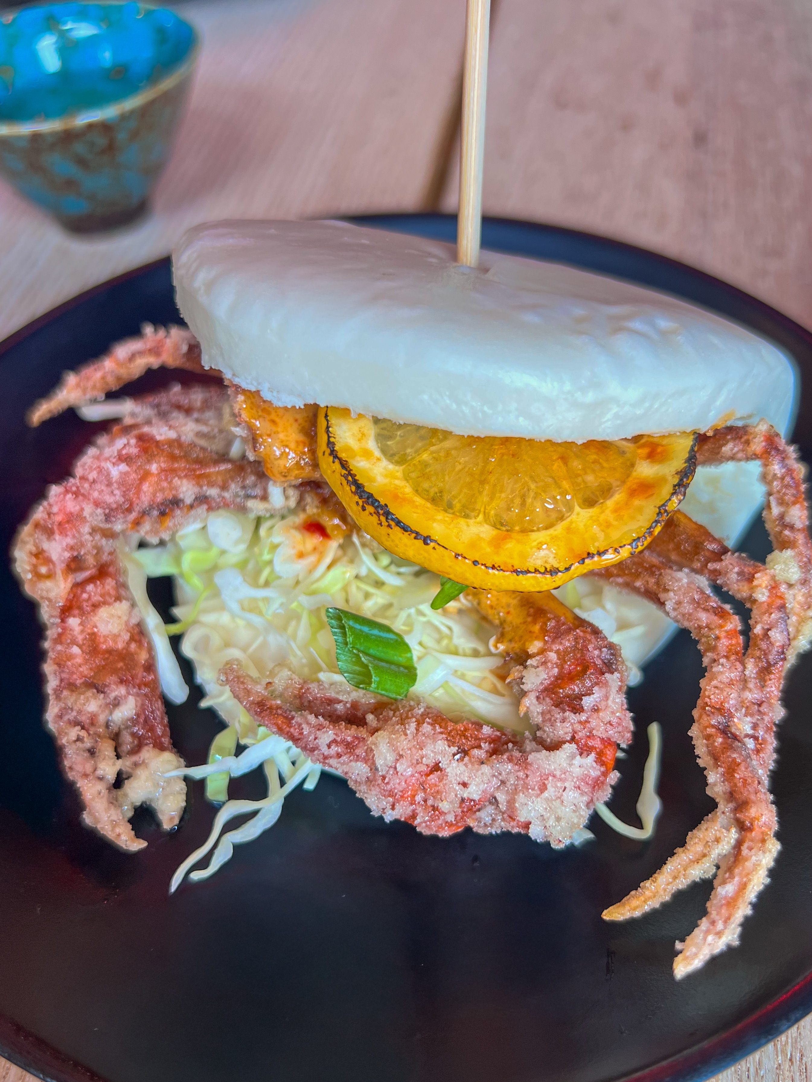 Whole fried soft-shell crab, bright orange and crispy, artfully placed inside a soft white bao bun with shredded lettuce